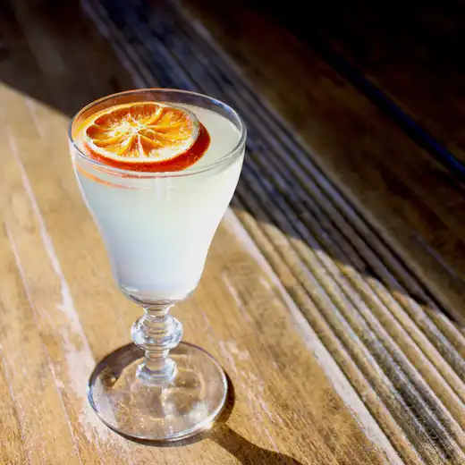 Pale cocktail with a dried orange slice garnish in a stemmed glass on a wooden table