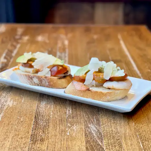 Two crostini topped with shrimp, avocado, and shaved cheese on a white plate