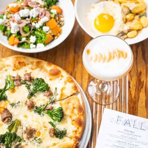 Pizza with sausage, side salad, bowl with a fried egg, and a foam-topped cocktail